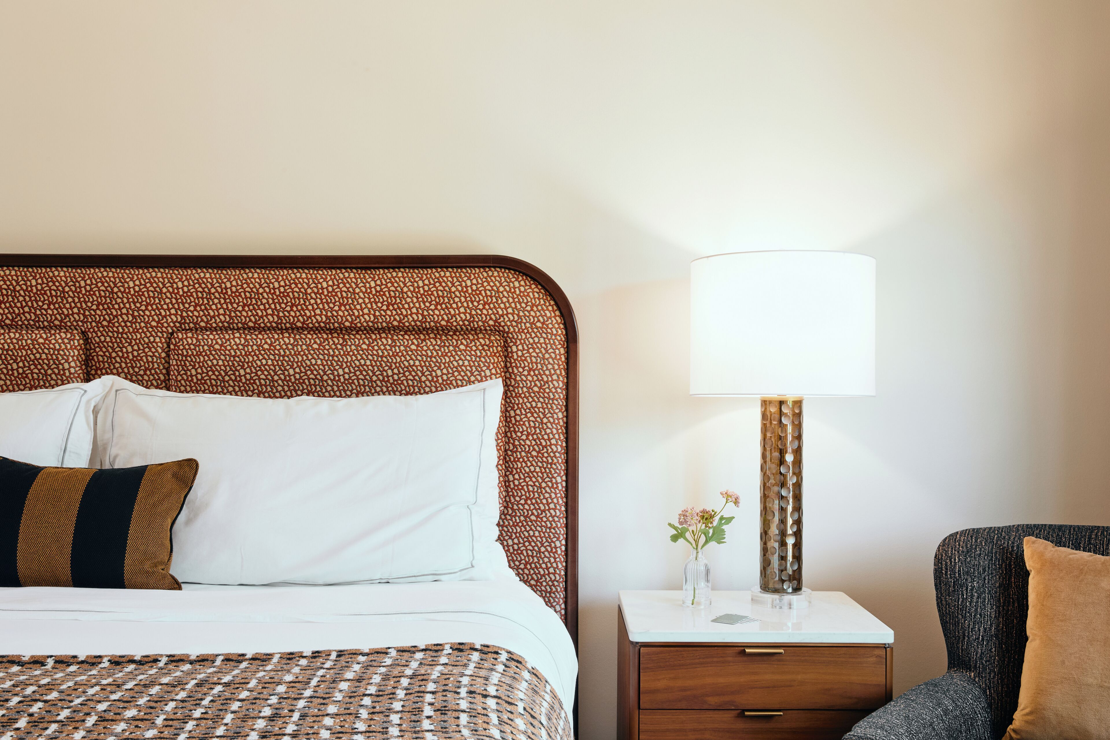 Een netjes opgemaakt bed met een beddengoed met patroon, een nachtkastje met een lamp en een stoel met kussen.
