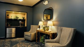 Una habitación con dos sillones, una mesita con lámparas, un espejo y un estante empotrado con plantas y un refrigerador.