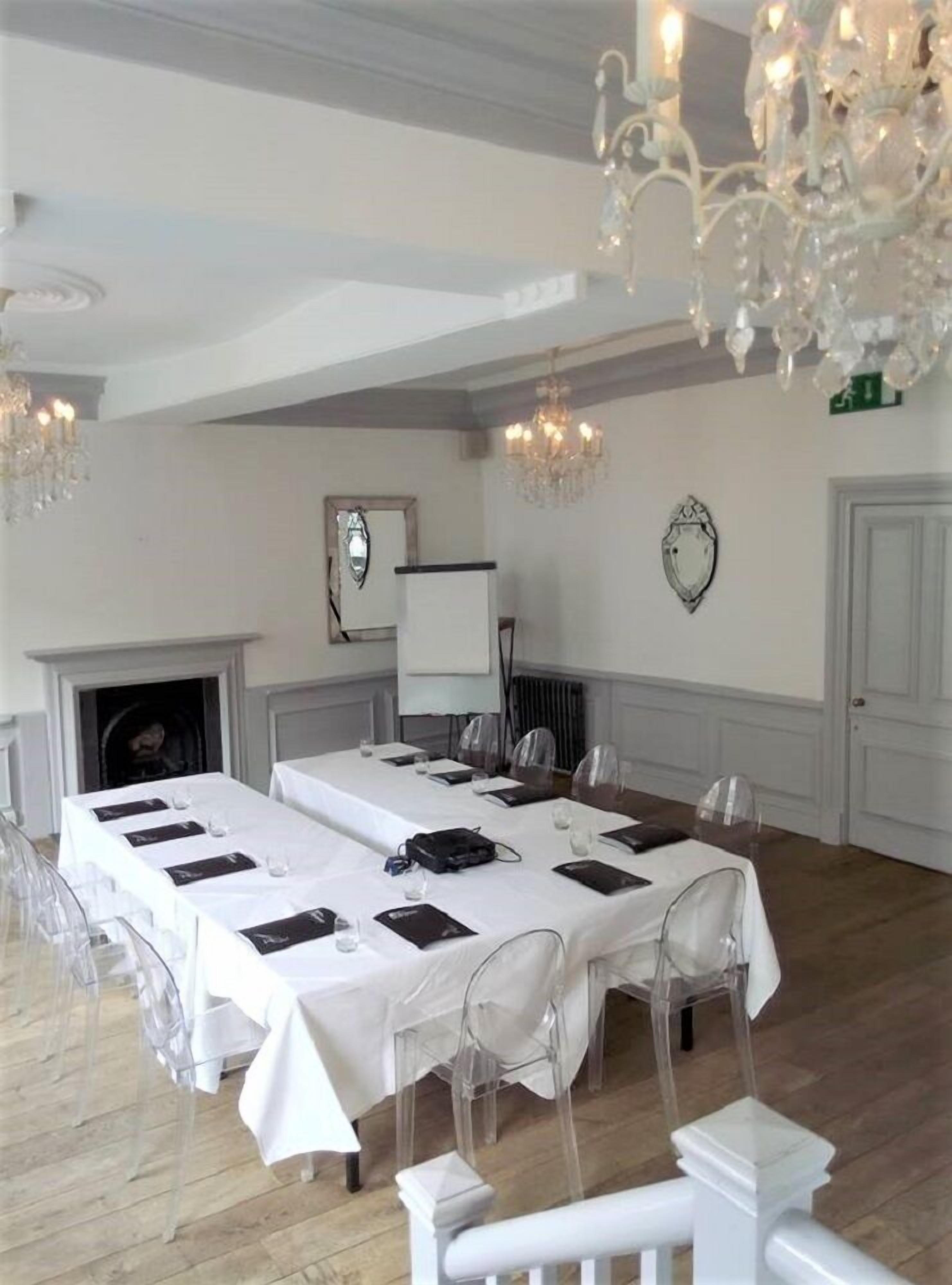 A conference room with a long table set for meetings, clear chairs, a fireplace, and chandeliers.