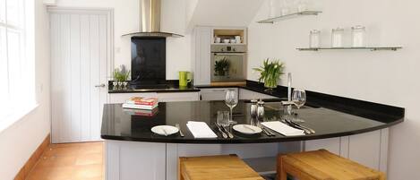 A modern kitchen with a black countertop, wooden stools, and glass shelves above.