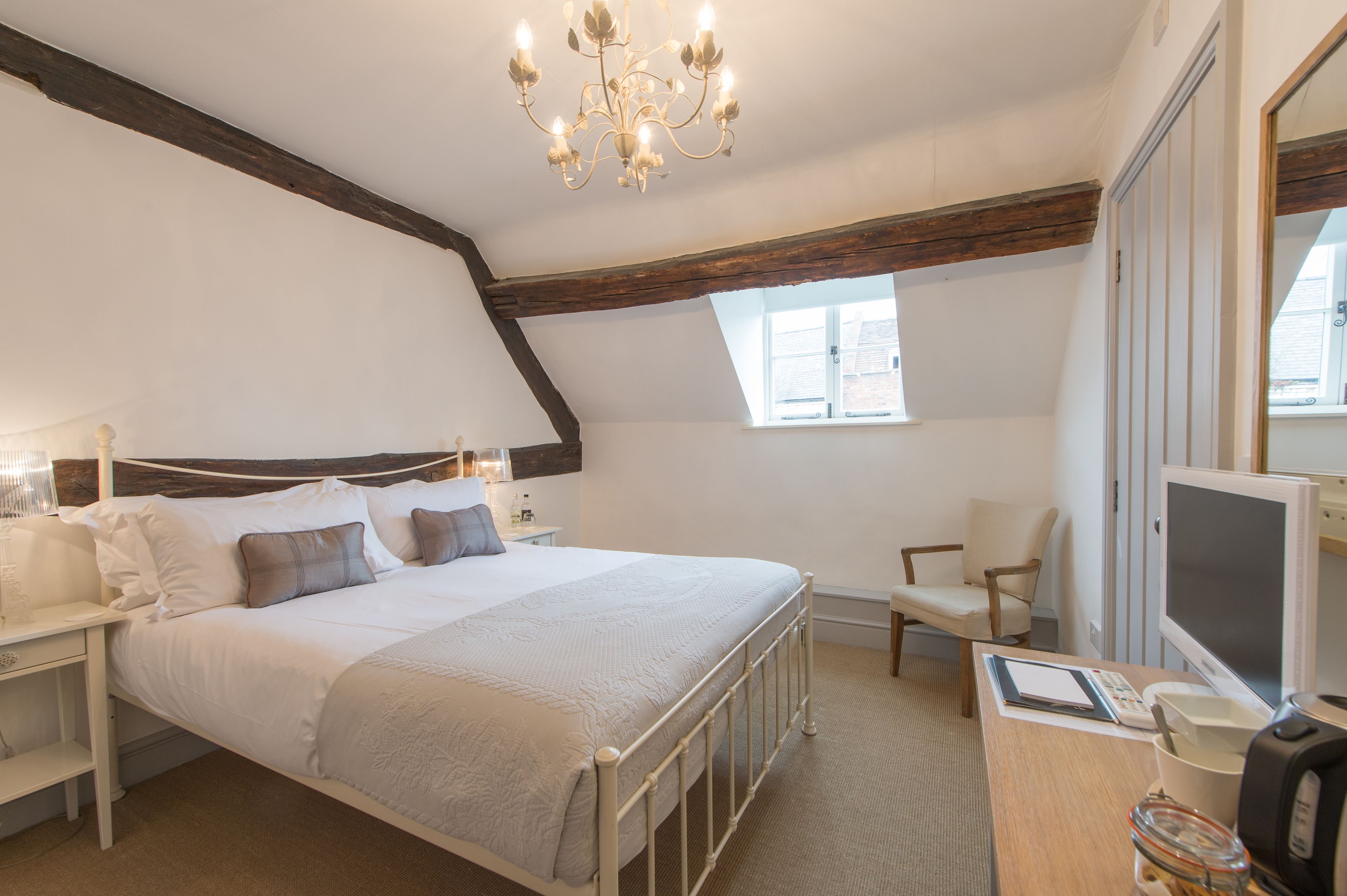 A bedroom with a large bed, a desk with a computer, a chair, a window, and a chandelier.