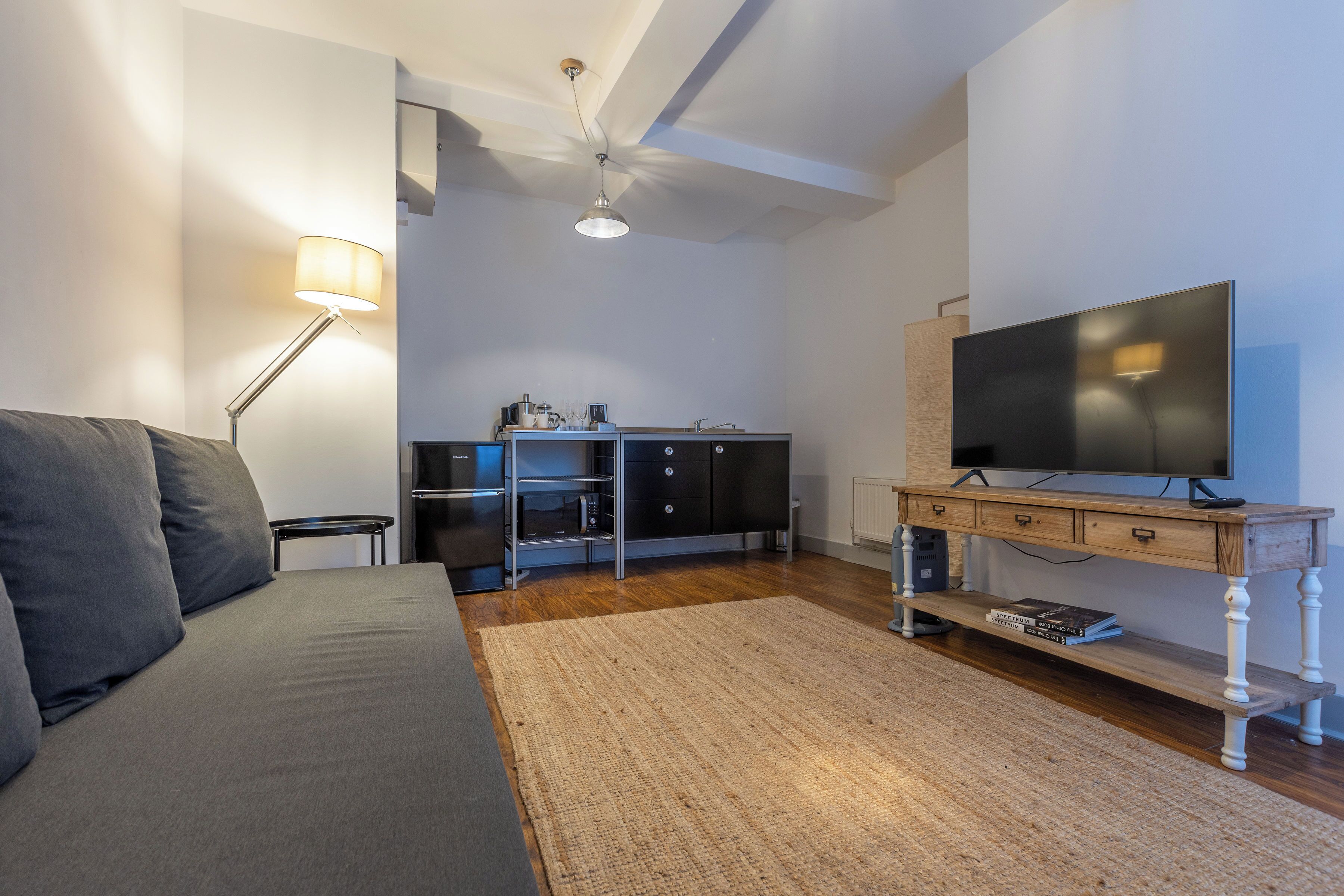 A modern living room with a sofa, a TV on a wooden stand, a lamp, and a small table with a coffee machine.