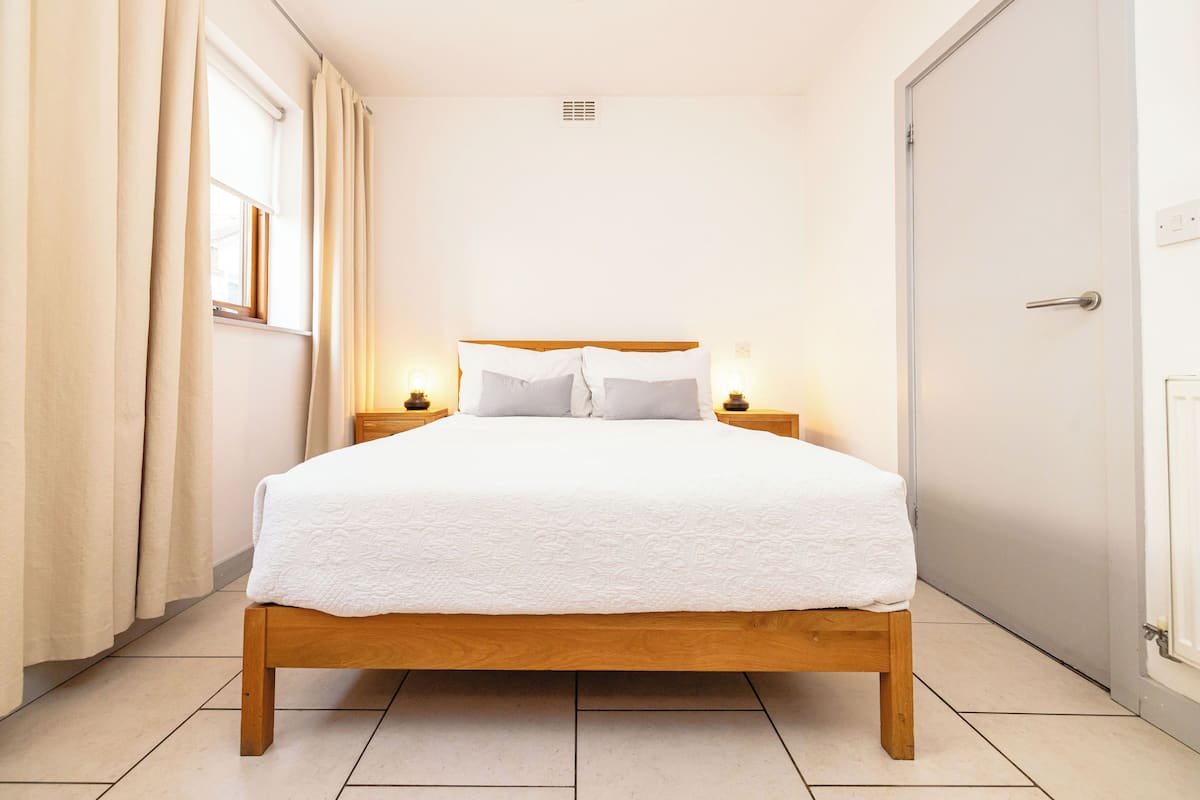 Un dormitorio con cama de madera, ropa de cama blanca, dos lámparas de noche y una ventana con cortinas.