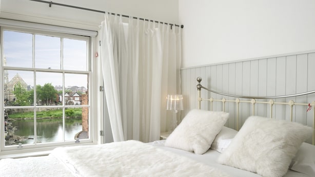 A bedroom with a large window, a bed with white bedding, and a view of a river and buildings. - Lion and Pheasant Hotel (Shrewsbury)