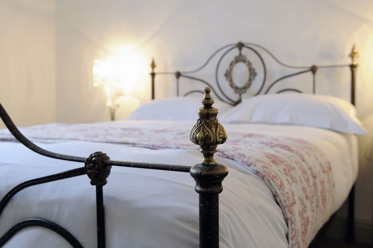 Una cama con estructura metálica y un cabecero decorado, una colcha blanca y una lámpara de noche.
