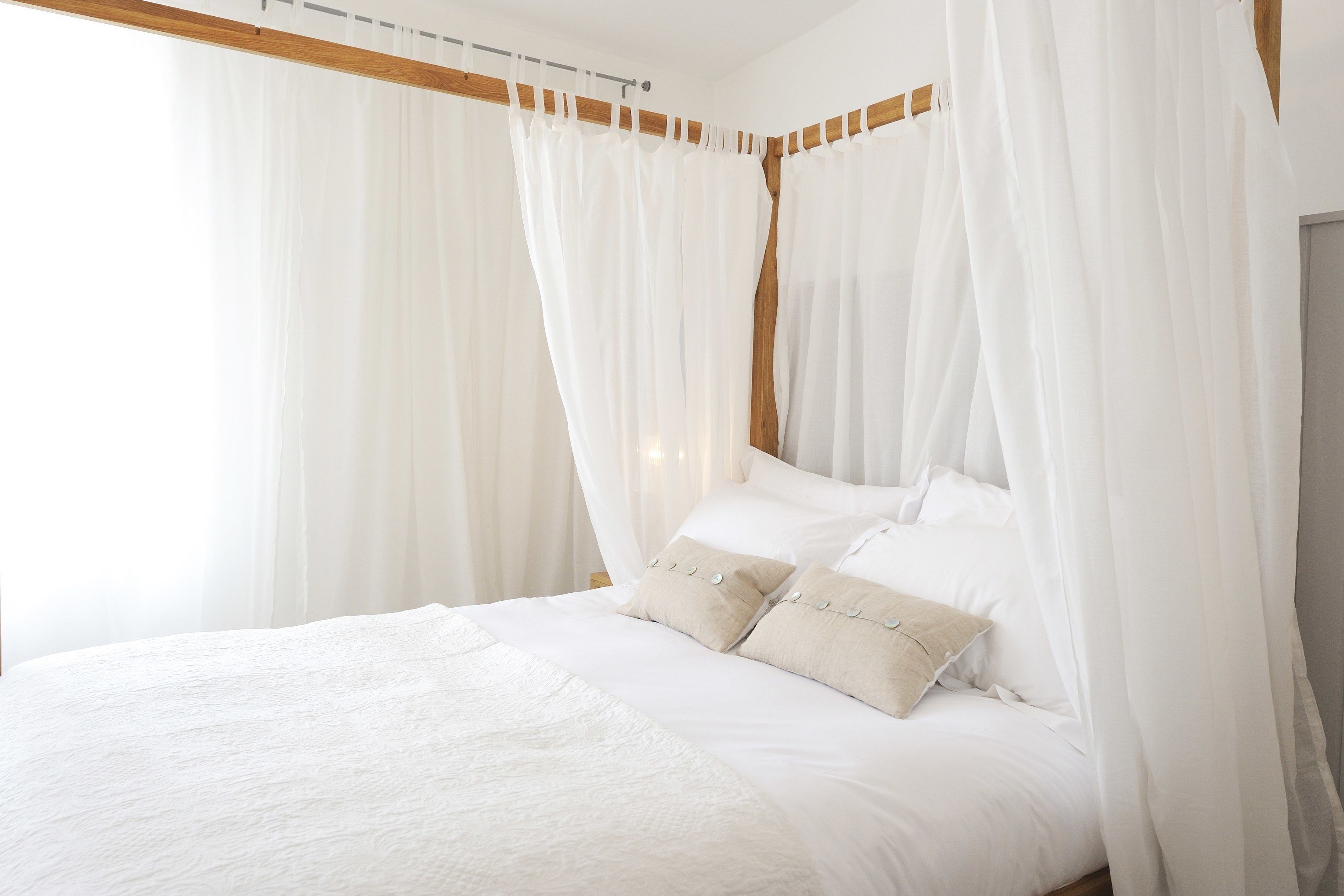 A bed with white bedding and a canopy, a wooden headboard, and a window with sheer curtains.