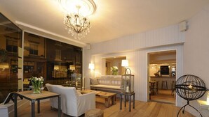 A modern living room with a chandelier, a sofa, a coffee table, and a wicker ottoman. There is a glass door leading to a kitchen area. - Lion and Pheasant Hotel (Shrewsbury)