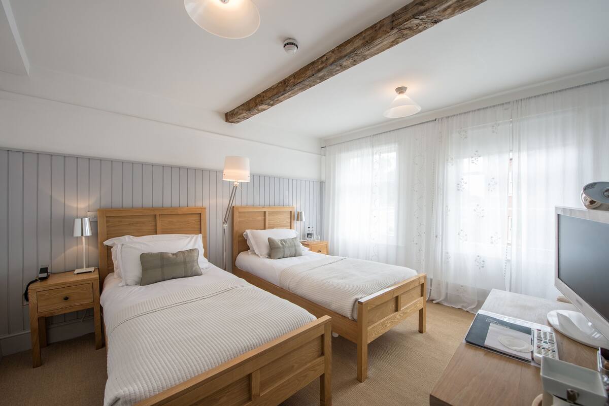 Habitación de hotel con dos camas de madera, un escritorio con ordenador y una ventana con cortinas translúcidas.