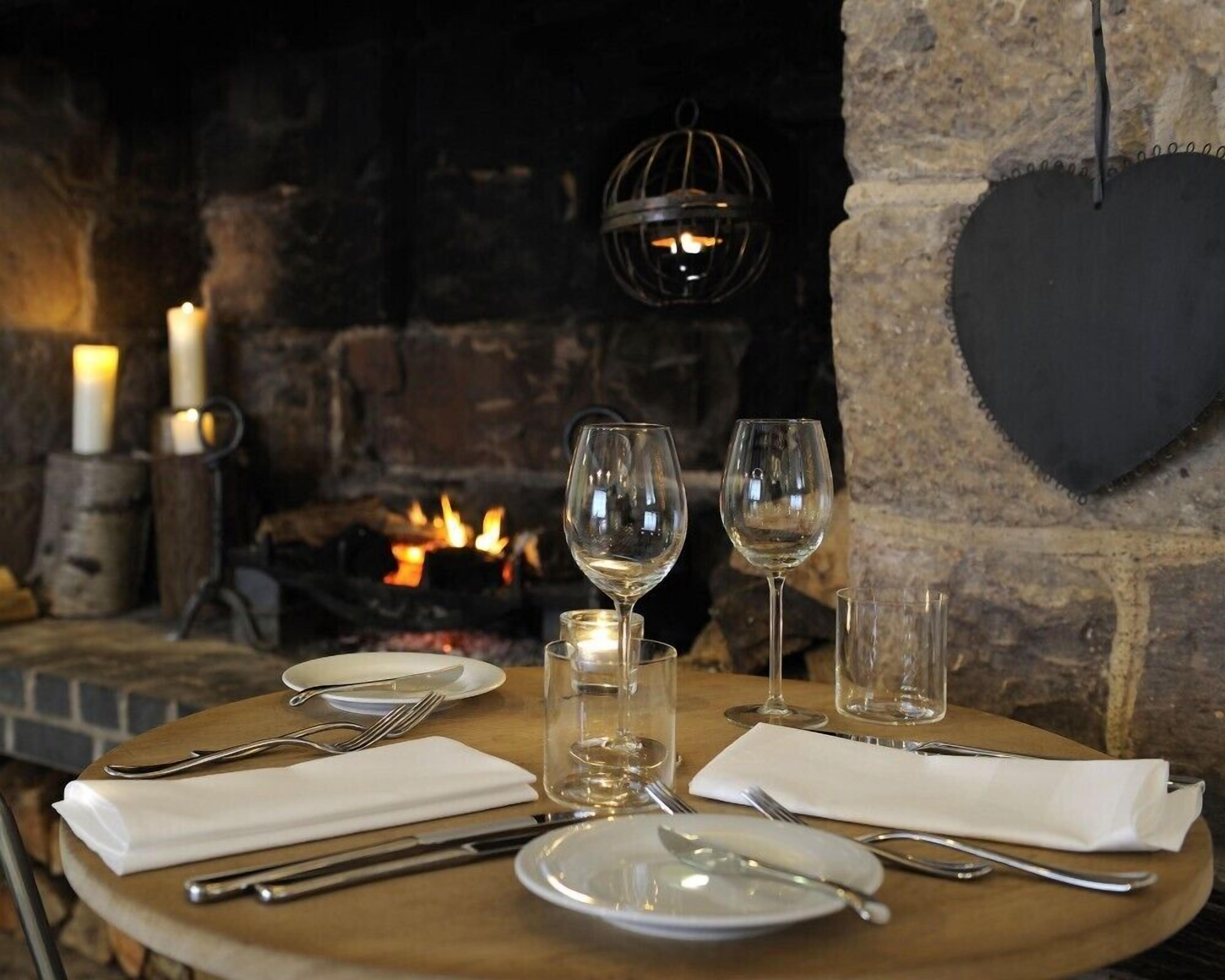 A table set for two with wine glasses, plates, and cutlery, a candle, and a fireplace in the background.