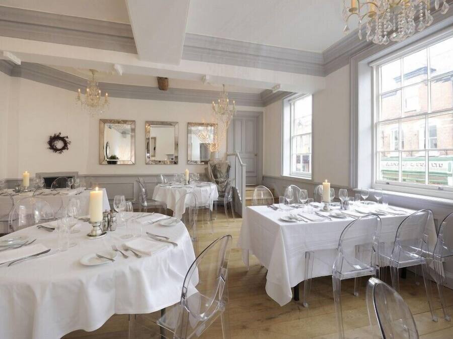 A dining area with round tables set for six, clear chairs, white tablecloths, candles, and chandeliers.