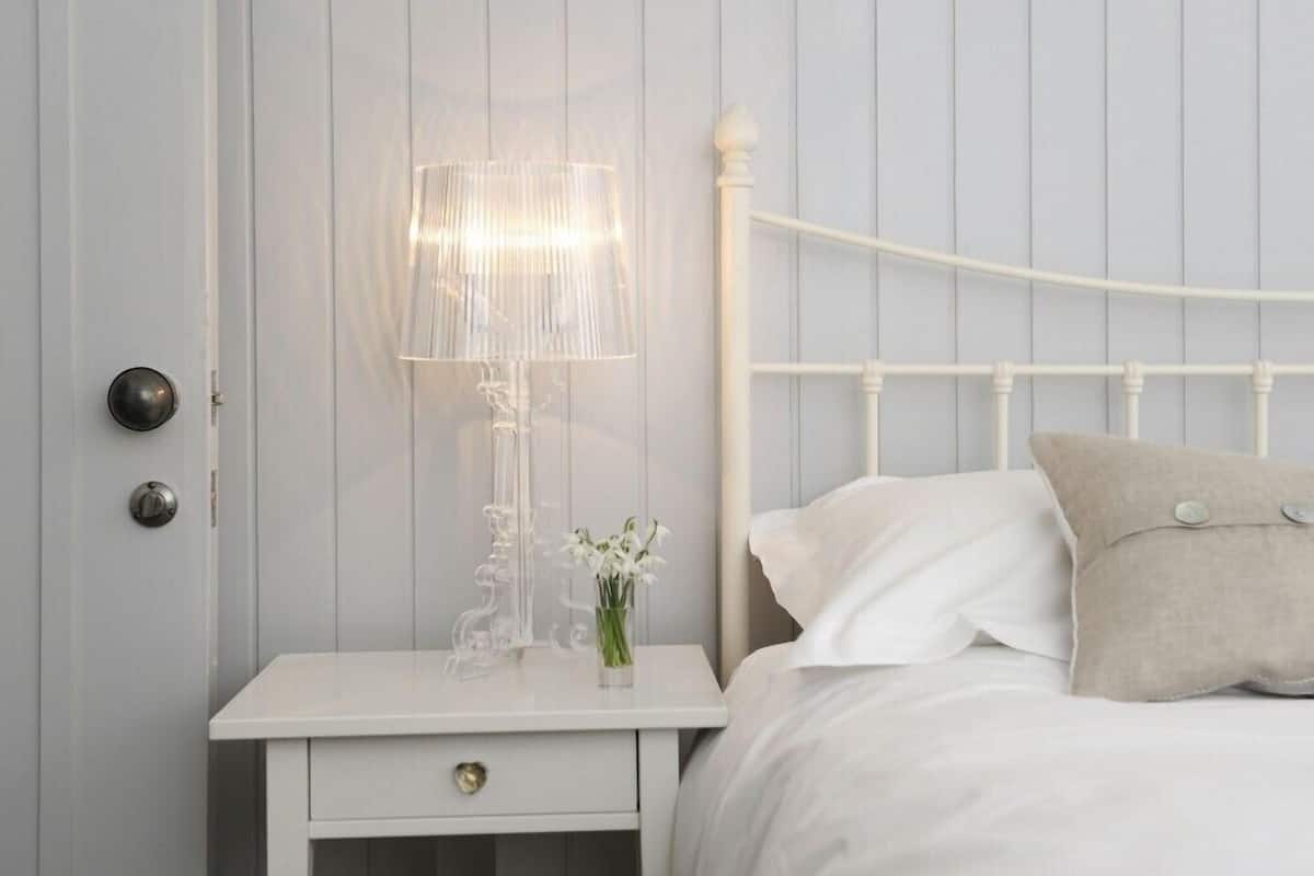 Un dormitorio con una cama blanca, una mesita de noche con lámpara y una puerta con pomo redondo.