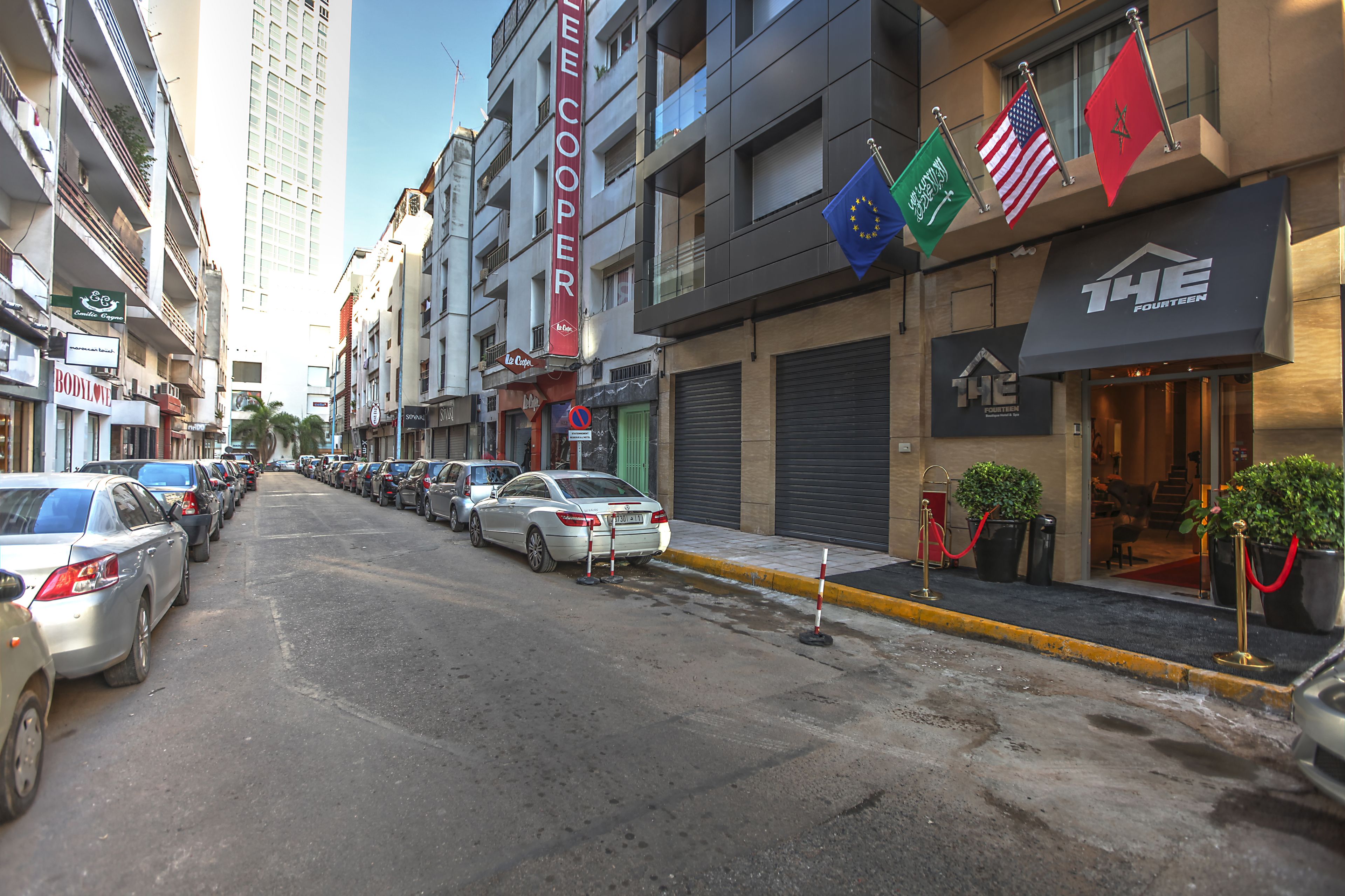 Une rue bordée de voitures garées, un immeuble orné de drapeaux et un magasin fermé dont l’enseigne indique « THE FOURTEEN ».