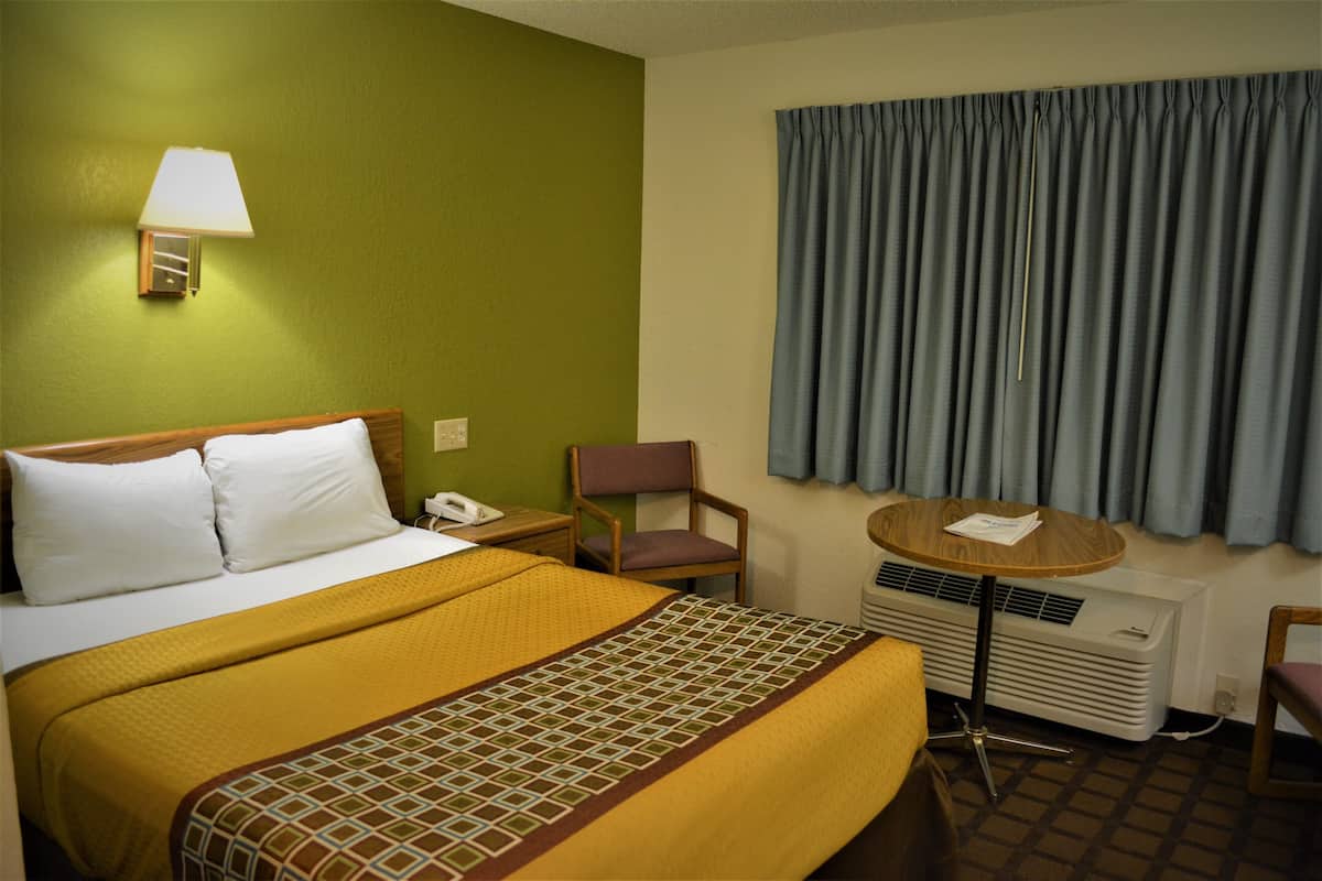 Ein Hotelzimmer mit Bett, Stuhl, kleinem Tisch, Lampe und einem Fenster mit Vorhängen.