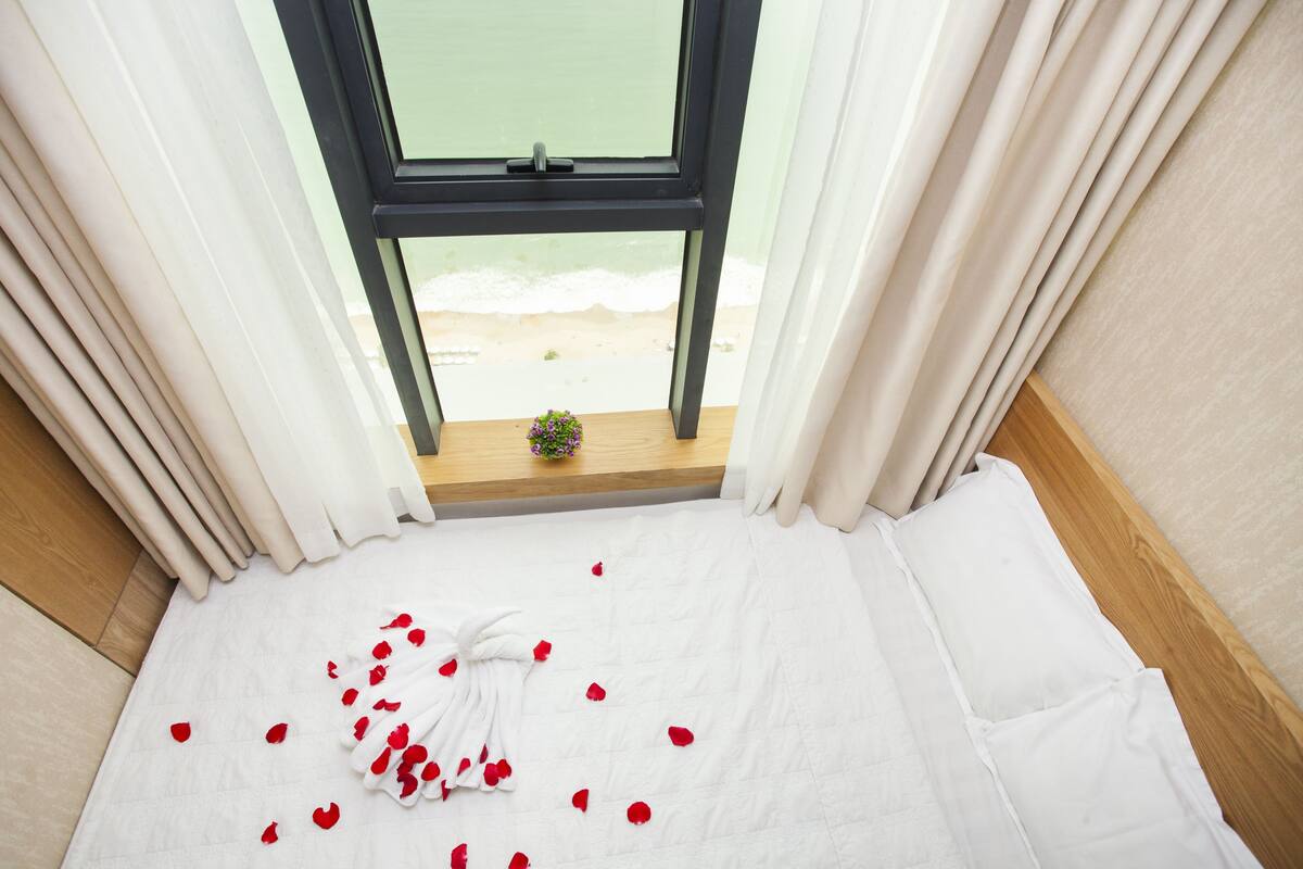A bed with white bedding, a small potted plant, and red rose petals on the floor.