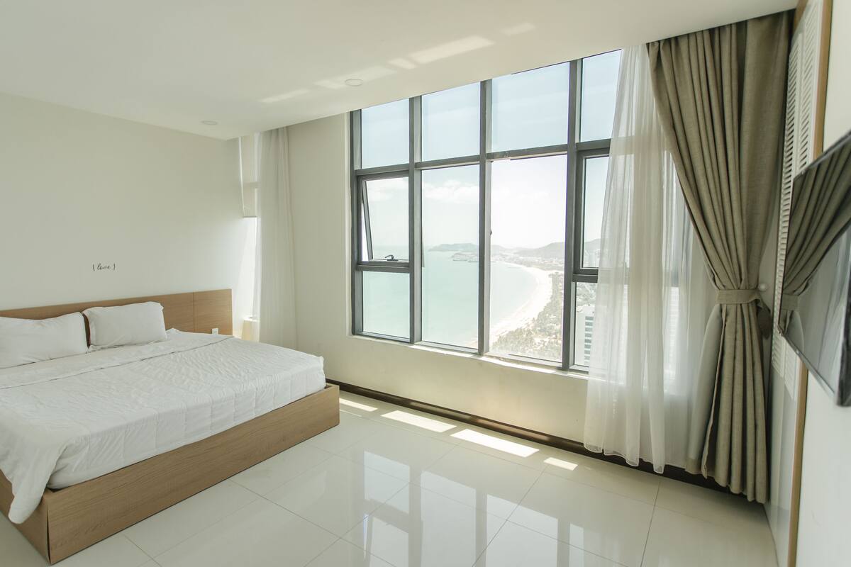 A modern hotel room with a large bed, a view of the sea, and a large window with curtains.