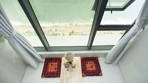 A room with a large window overlooking a beach, a small table with a plant, and two red patterned mats. - Beach Front Apartment (Nha Trang)