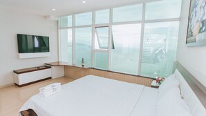 A modern hotel room with a large bed, a flat-screen TV mounted on the wall, a bedside table with a bottle, and a large window offering a view of the city. - Beach Front Apartment (Nha Trang)