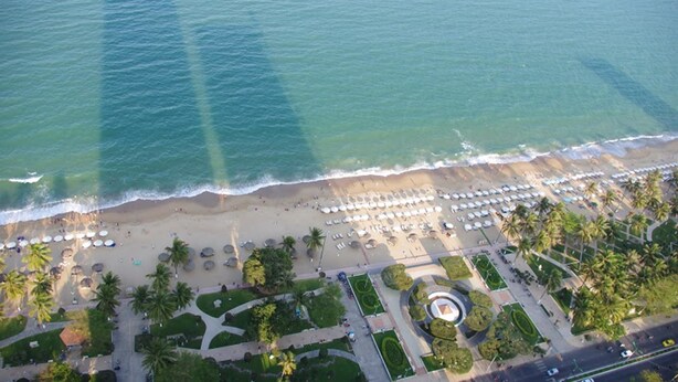 An aerial view of a beach with rows of umbrellas and palm trees, a parking lot, and a landscaped area with pathways and greenery. - Beach Front Apartment (Nha Trang)