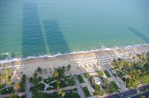 Vista aerea di una spiaggia con file di ombrelloni e palme, un parcheggio e un'area verde con vialetti e vegetazione.