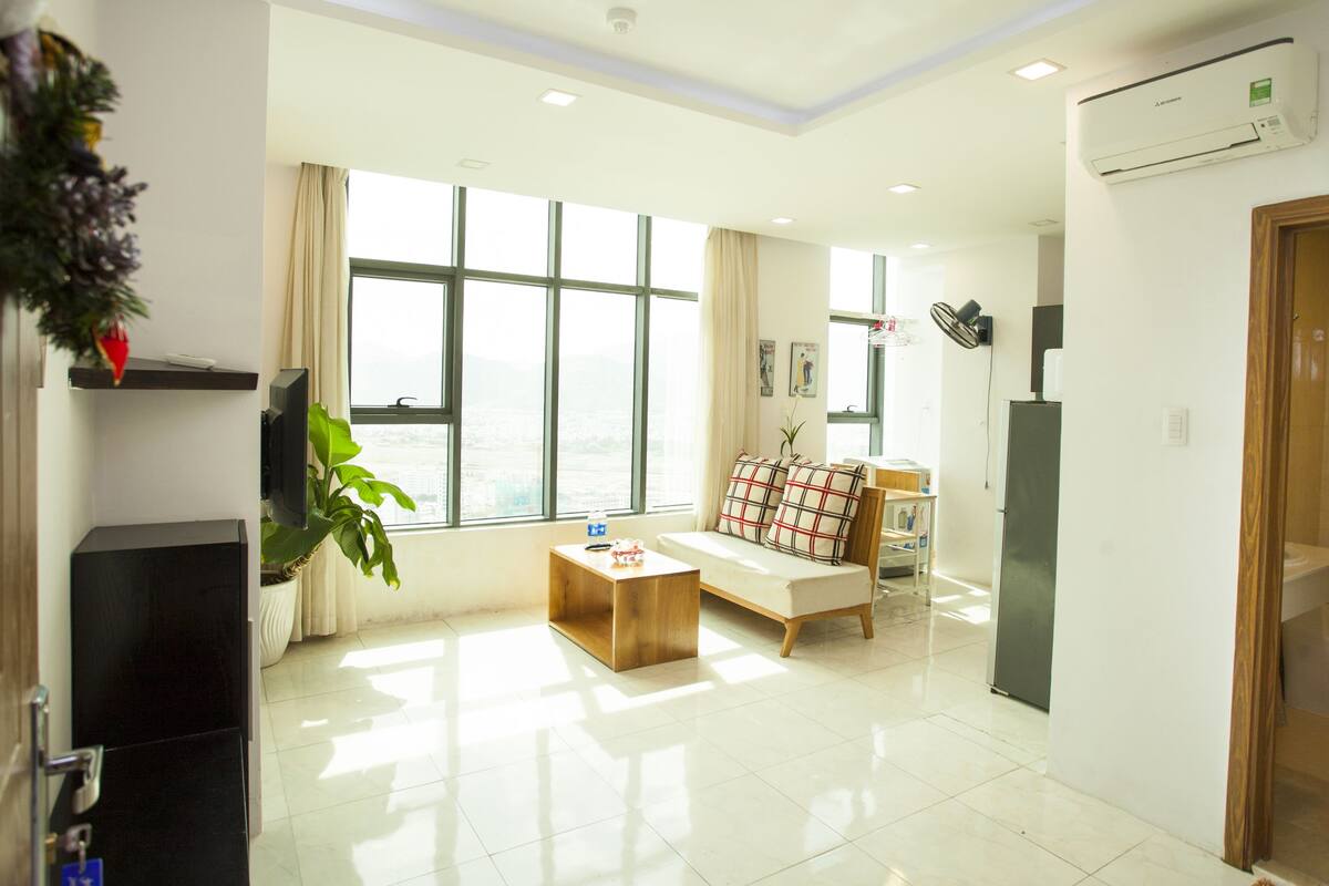 A modern living room with a sofa, coffee table, and a TV. There is a large window with curtains, a plant, and a view of the city.