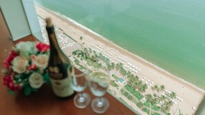 A table with a bottle of champagne and two wine glasses, overlooking a beach with umbrellas and palm trees. - Beach Front Apartment (Nha Trang)