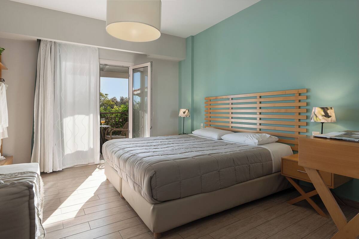 A bedroom with a bed, a desk, and a chair. There is a window with curtains, and a door leading to an outdoor area.