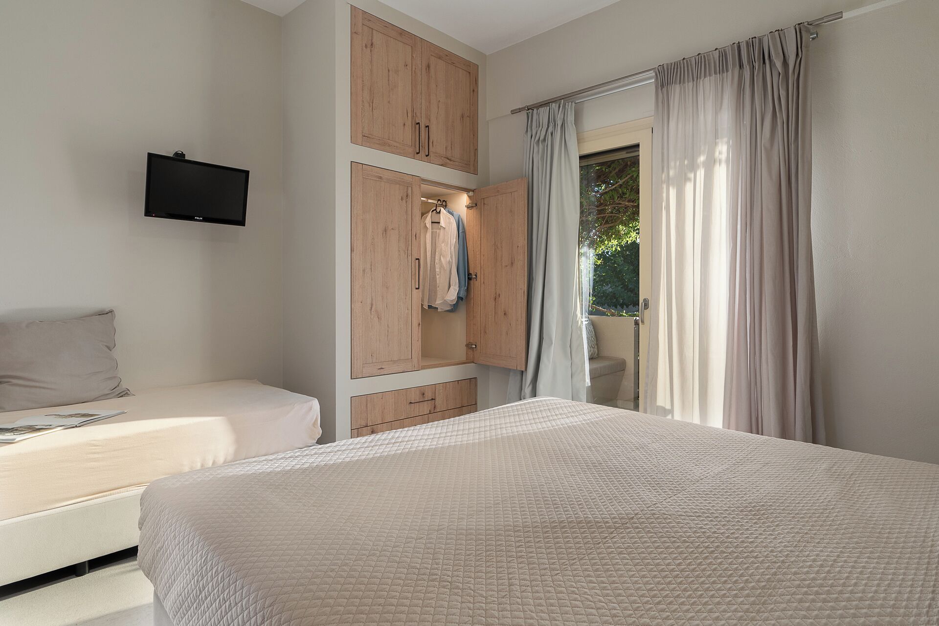 A bedroom with a bed, a built-in wardrobe, a TV mounted on the wall, and a window with curtains.