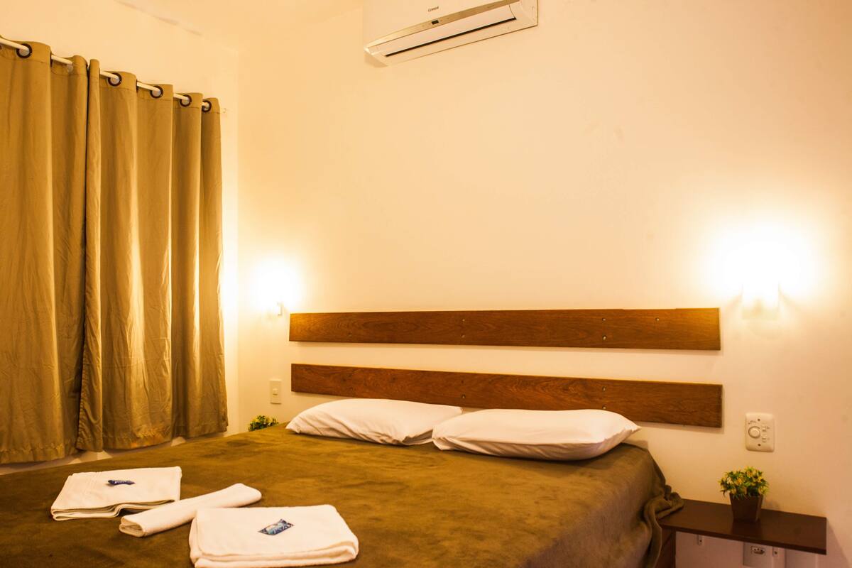 A hotel room with a bed, two pillows, a headboard, a nightstand, a plant, and a curtain.