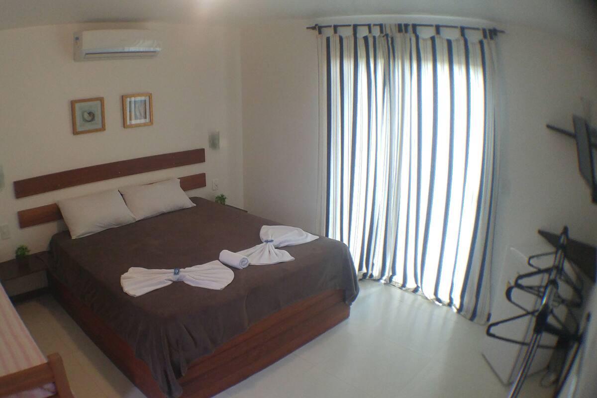 A hotel room with a bed, a desk, a chair, a window with curtains, and an air conditioning unit.