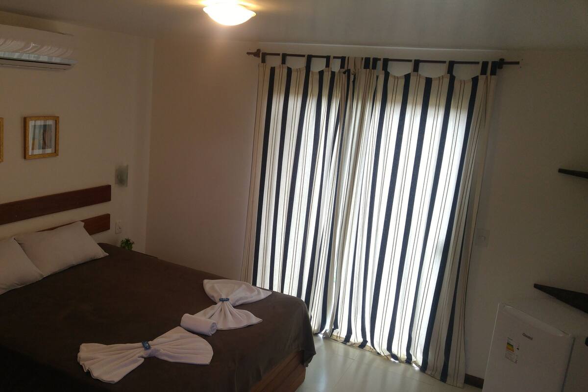 A hotel room with a bed, a wall-mounted air conditioner, two framed pictures, and a window with striped curtains.