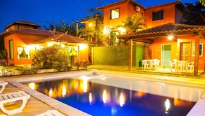 Une piscine avec des chaises longues, un patio couvert avec des fauteuils et une maison avec un toit en tuiles et un éclairage chaleureux.