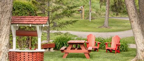 A picnic setup with a wooden table and two red Adirondack chairs, surrounded by trees and a grassy area.