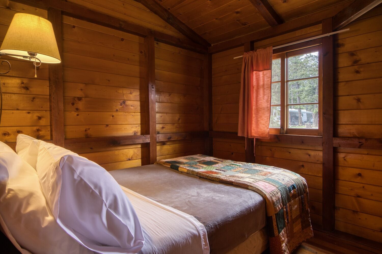 Une pièce de style cabane en bois, avec un lit, une fenêtre avec des rideaux et une lampe fixée au mur.