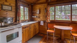 A cabin interior with wooden walls, a kitchen with a stove and sink, a dining table with two chairs, and a window with a view of trees and a house.