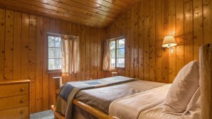A wooden cabin bedroom with a bed, two windows, a nightstand, and a lamp.
