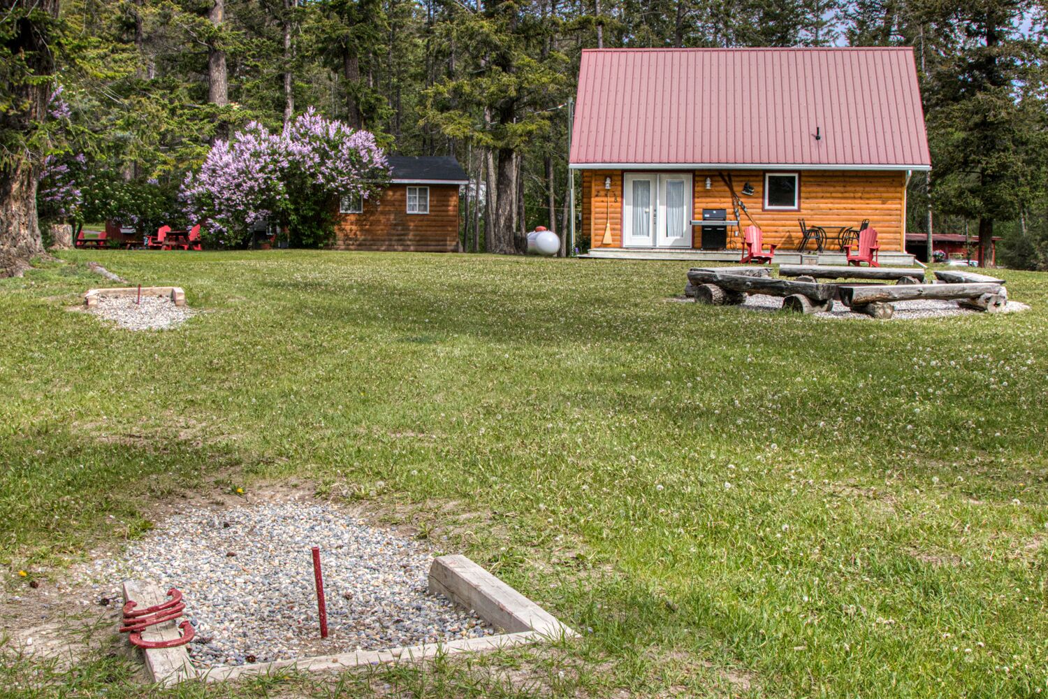 A wooden cabin with a red roof, surrounded by a grassy area with a small gravel path and a wooden bench.