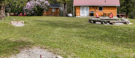 A wooden cabin with a red roof, surrounded by a grassy area with a small gravel path and a wooden bench.