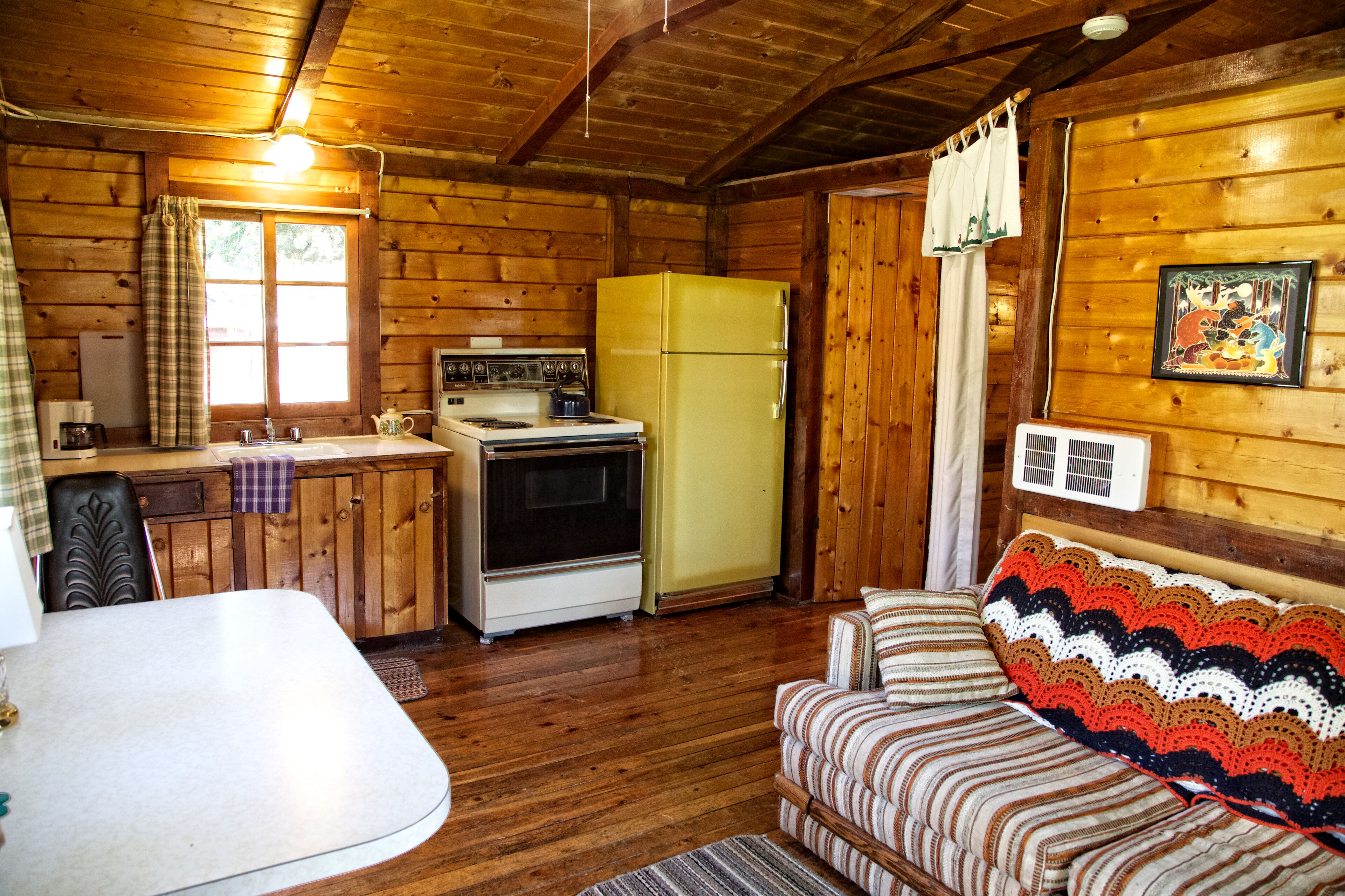 A wooden cabin interior with a kitchen, dining area, and a cozy living space with a couch and a crocheted throw.