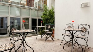 Uma área de pátio ao ar livre com quatro mesas redondas e cadeiras, um vaso de planta e uma porta de vidro que dá para um edifício.