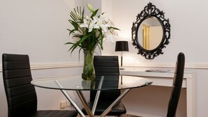 Uma sala de jantar com uma mesa de vidro, cadeiras pretas, um vaso com flores, um abajur preto e um espelho na parede.