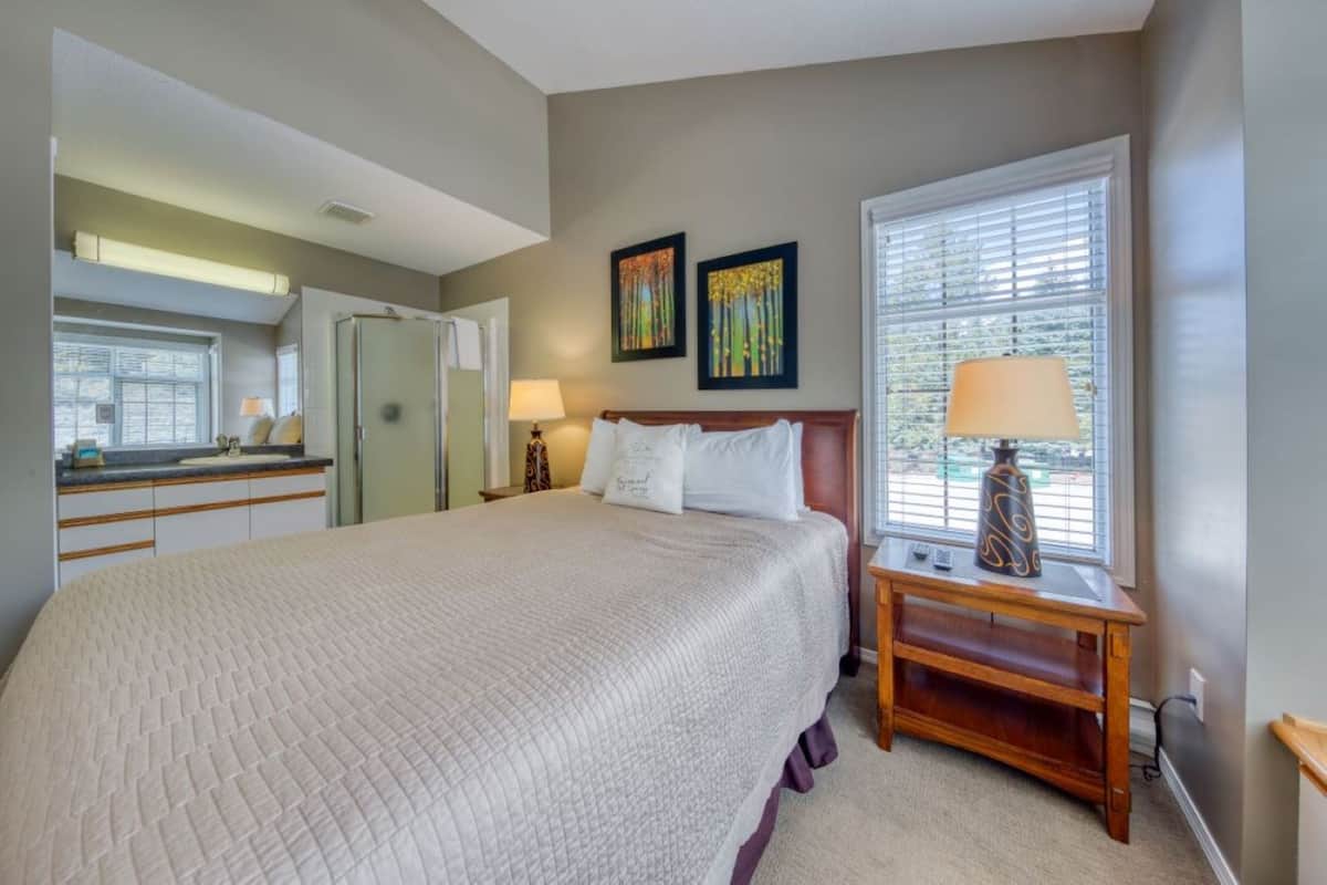 A bedroom with a large bed, a nightstand, a lamp, and a window with blinds.