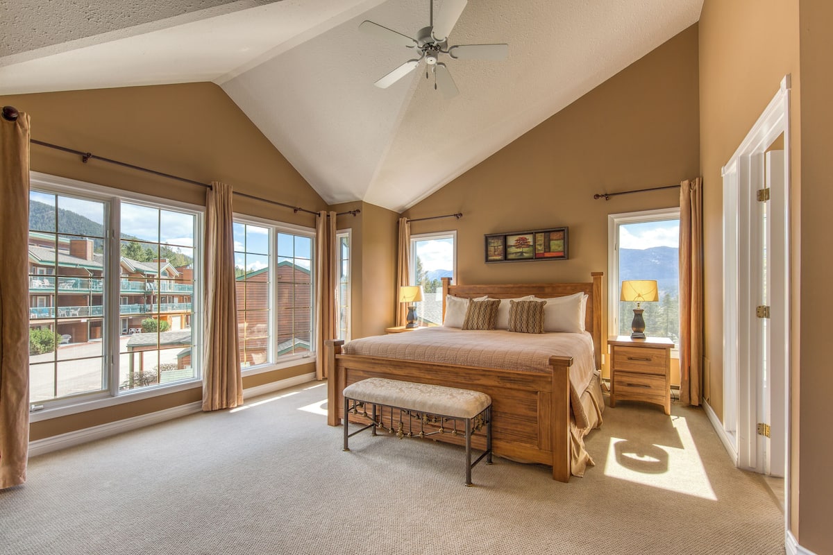 A spacious bedroom with a large bed, a bench, and a view of a mountain and a building through large windows.