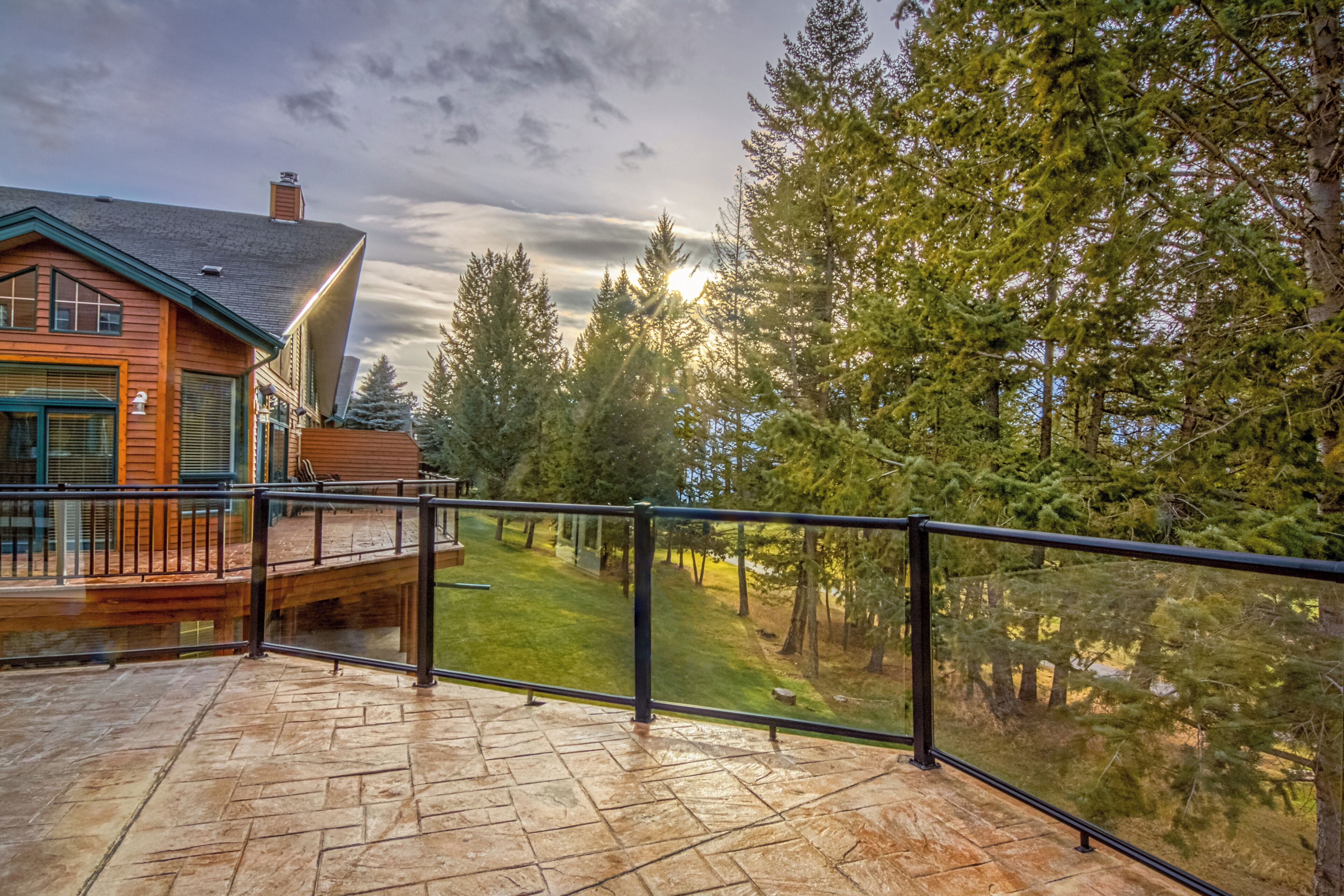 A wooden cabin with a stone deck and a metal railing, surrounded by trees and a clear sky.