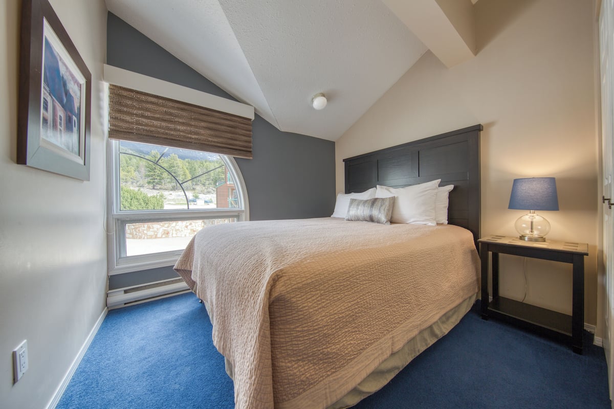 A bedroom with a large bed, a nightstand with a lamp, a window with a view of greenery, and a framed picture on the wall.