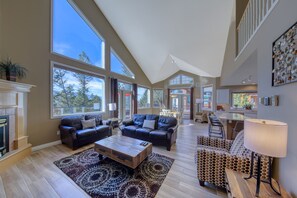 A spacious living room with a fireplace, large windows, and a high ceiling.
