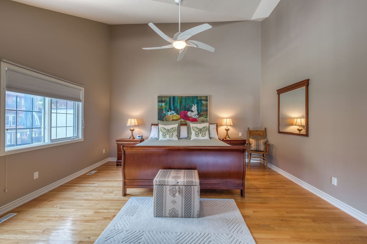 A bedroom with a large bed, a ceiling fan, a window with blinds, a painting on the wall, and a nightstand with a lamp.