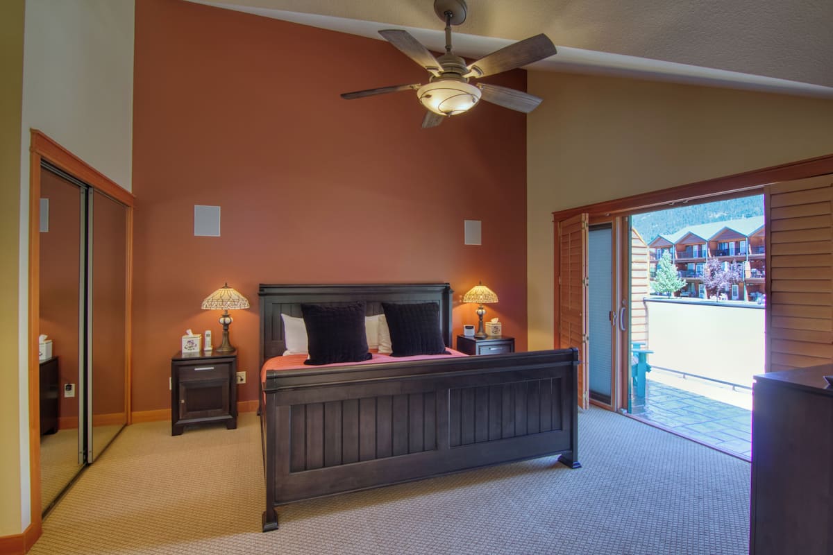 A bedroom with a large bed, two bedside tables with lamps, a ceiling fan, and a view of a building through an open door.