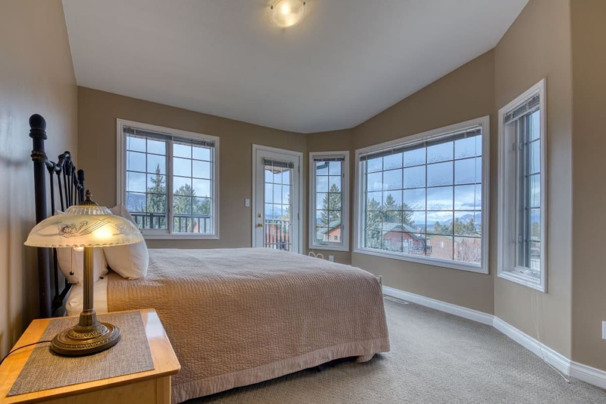 A bedroom with a large bed, a nightstand, a lamp, and multiple windows offering a view of the outside.