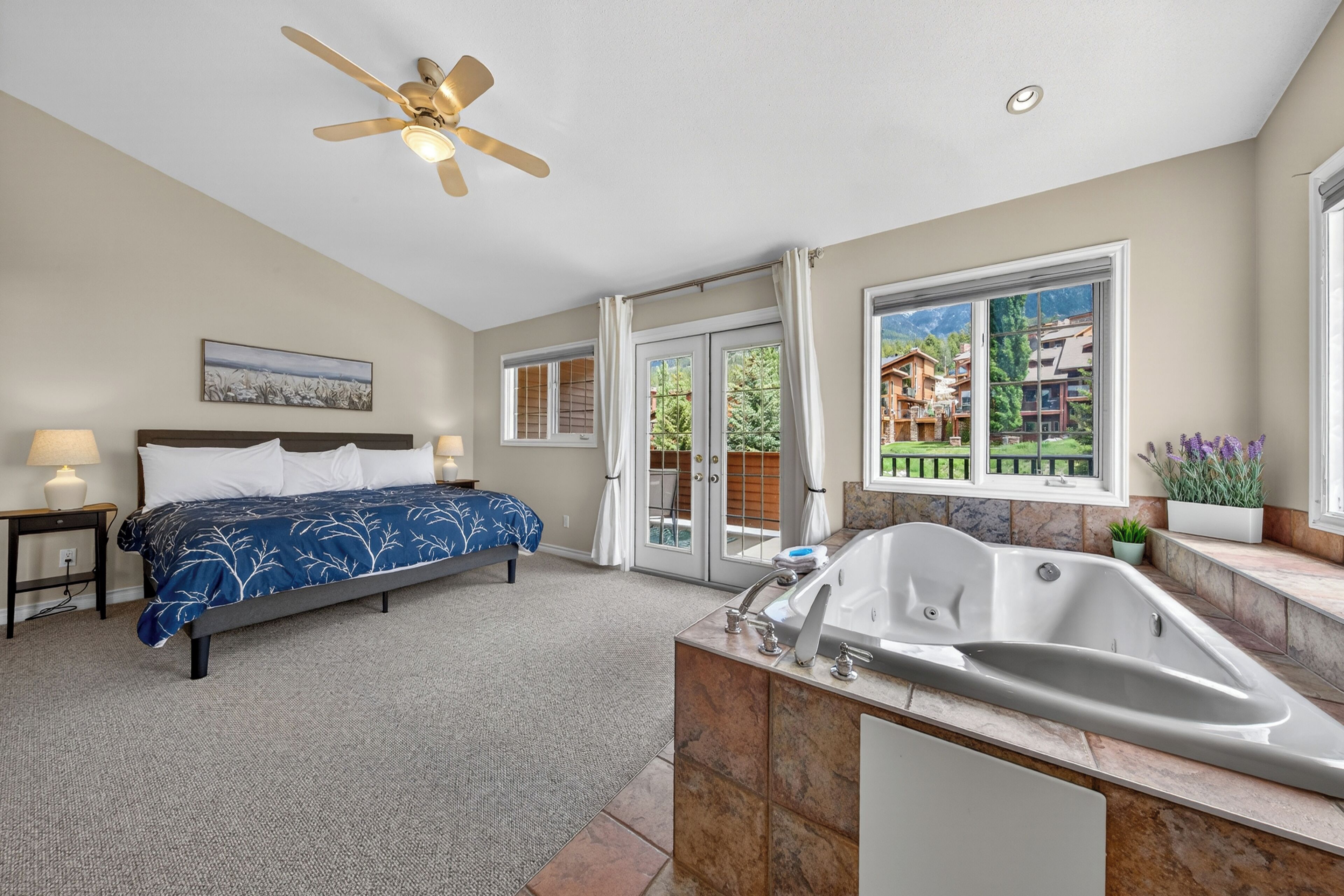 A bedroom with a bed, a ceiling fan, a large window with curtains, and a bathroom with a bathtub and sink.