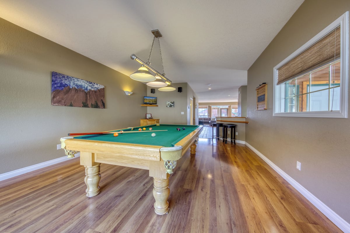 A pool table in a room with wooden floors, a ceiling light fixture, and a window with blinds.